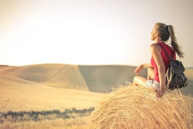 Woman with a backpack sitting on a hay bale, enjoying a serene sunset over a vast countryside field.